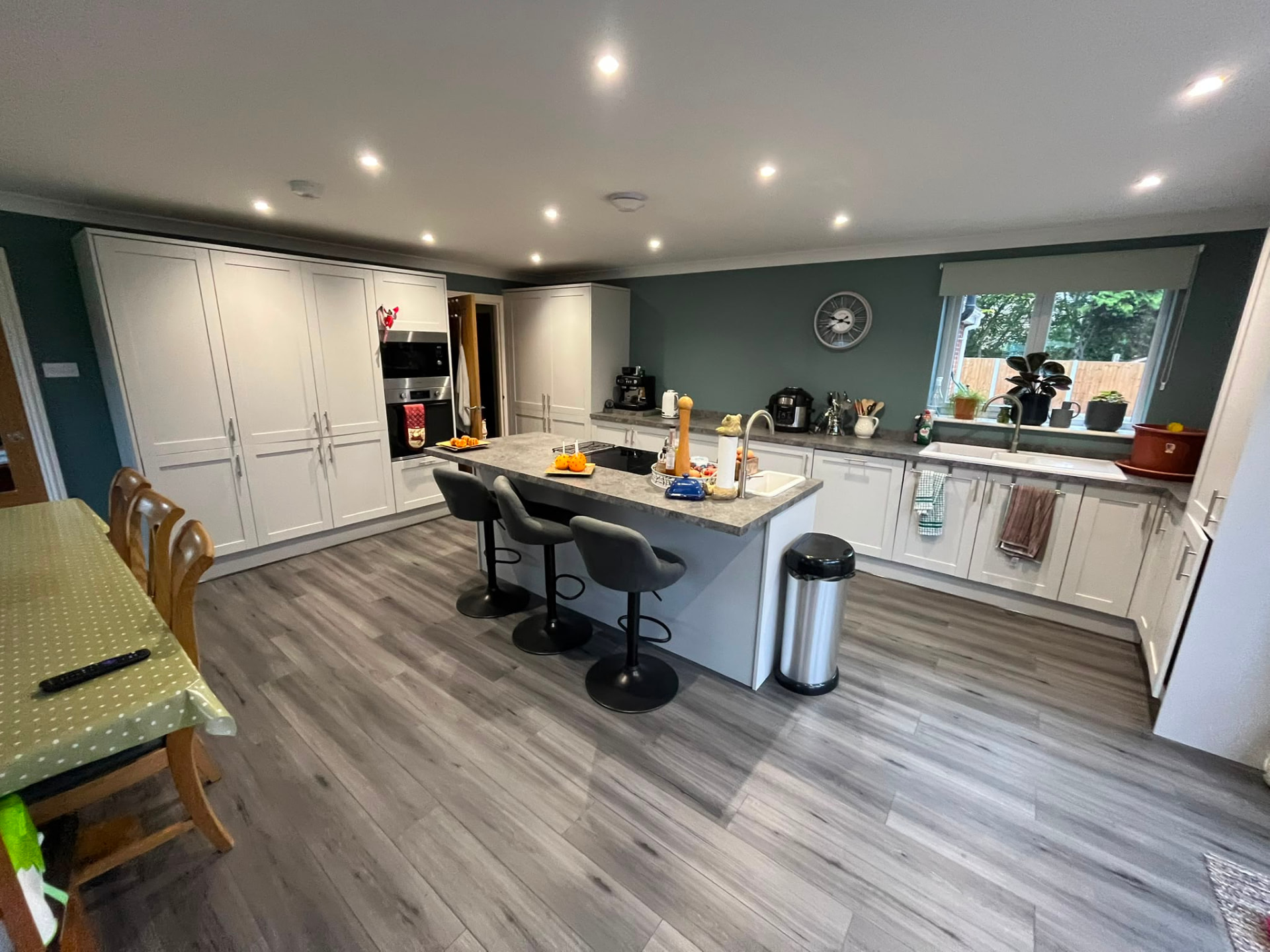Open-plan kitchen with pale shaker cabinetry, a stone-effect island with three bar stools and deep green walls
